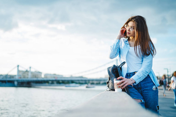 Charming young woman dressed in casual wear calling on smartphone using roaming connection and enjoying beauty of river in modern city.Attractive tourist talking on mobile phone in urban setting - Fotó, kép
