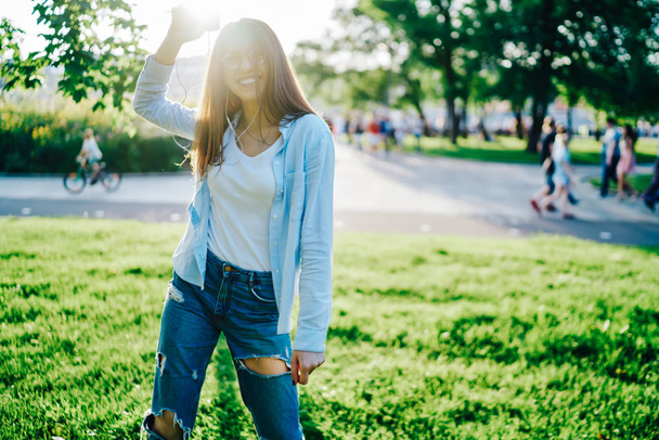 Portrait of cheerful hipster girl dressed in casual wear enjoying music in earphones connected to smartphone in urban setting in sunny park.Positive young woman smiling at camera while listening audio - Fotografie, Obrázek