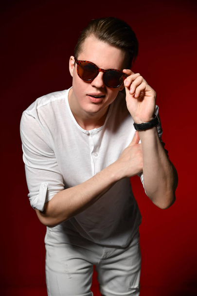 Gorgeous and stylish. Handsome young man in sunglasses keeping hands clasped and looking away while sitting on wooden chest against red background - Фото, зображення