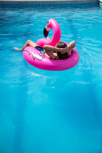 junger Mann schwimmt in einem aufblasbaren Flamingo in einem blauen Pool an einem sonnigen Sommerferientag - Foto, Bild