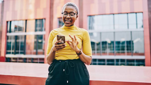 Happy african american woman excited with getting news about sales in web store on mobile phone, cheerful dark skinned hipster girl standing on urban background with smartphone laughing on conten - Foto, immagini