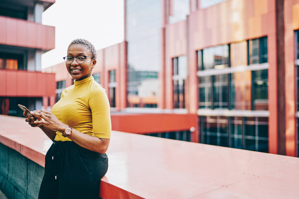 Portrait d'une Africaine américaine joyeuse qui se tient debout en milieu urbain à l'aide d'un téléphone intelligent et d'une connexion 4G en itinérance, une blogueuse à la peau foncée tenant un téléphone mobile pour bavarder - Photo, image