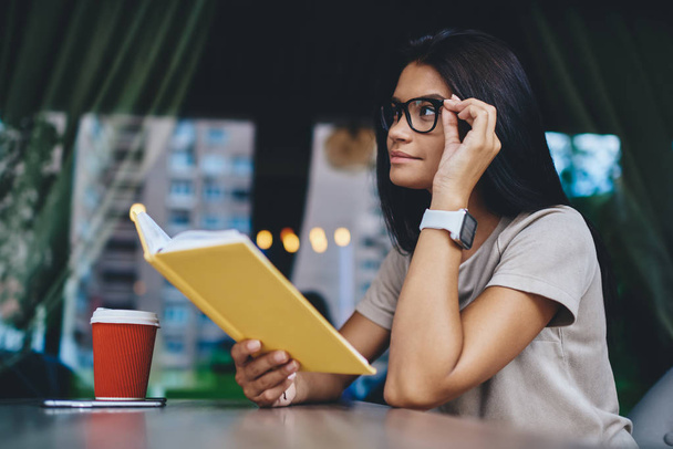 Pensive caucasian young woman in spectacles thinking about interesting novel plot sitting at cafe with coffee, dreamy hipster girl in eyewear analyzing literature spending free time on new book - Foto, imagen