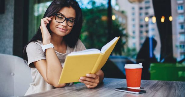 Positive hipster girl in spectacles for vision correction read new best seller during free time in cafe, young caucasian woman fond of literature satisfied with interesting book on coffee break - Photo, Image