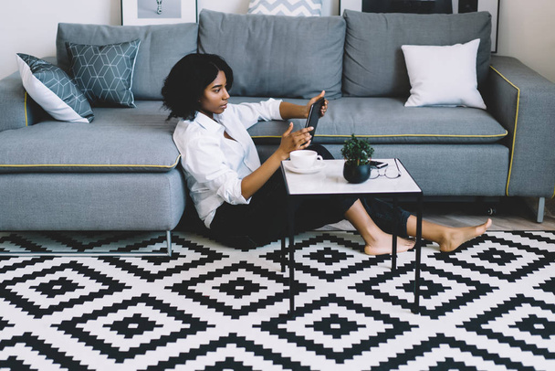 Африканська американська молода жінка, одягнена в повсякденне вбрання, сидить на підлозі в сучасній квартирі з стильним інтер'єром і меблями, дивлячись відео в блозі на сучасному сенсорному майданчику з використанням домашнього інтернет-підключення. - Фото, зображення