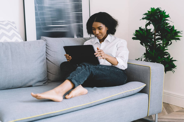 Pensive african american young woman watching live stream on website using home internet on digital tablet relaxing on couch in comfortable apartment.concentrated dark skinned female reading news - Zdjęcie, obraz