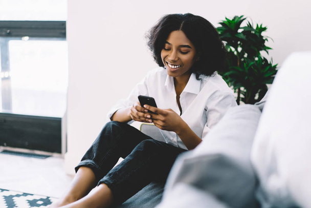 Cheerful dark skinned female blogger laughing during online chat with followers on phone using home internet.Positive african american young woman reading good news on smartphone relaxing on couch - Φωτογραφία, εικόνα