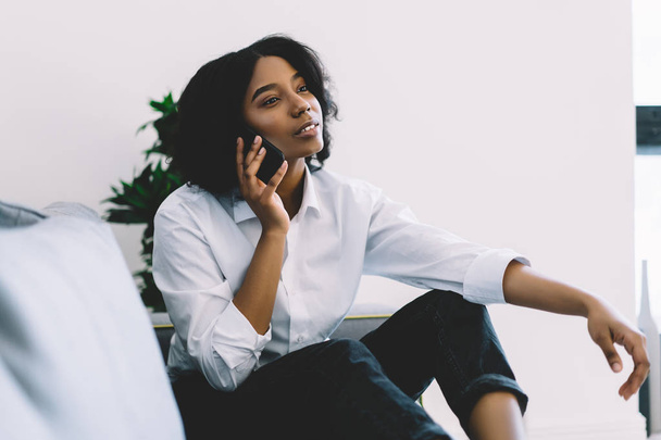 Beautiful african american hipster blogger talking with mobile operator on smartphone resting at home on cozy couch.Charming dark skinned young woman calling on mobile phone relaxing in flat - Photo, image