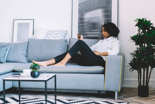 Stylowa, afrykańska młoda kobieta leżąca na wygodnej kanapie i czytająca powiadomienia w sieciach społecznościowych na smartfonie korzystając z domowego łącza internetowego.Kobieta blogger na czacie online przez telefon - Zdjęcie, obraz