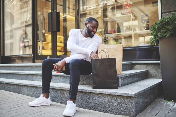 Smiling dark skinned hipster guy dressed in long sleeve shirt looking at shopping bags checking purchases, positive african american male happy about buying clothing in wear store with good discount - Fotografie, Obrázek