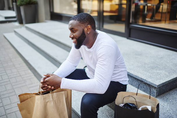Smiling afro american male customer in trendy wear sitting on stairs of store with bags, cheerful dark skinned hipster guy recreating on street publicity area after shopping and buying purchases - Φωτογραφία, εικόνα