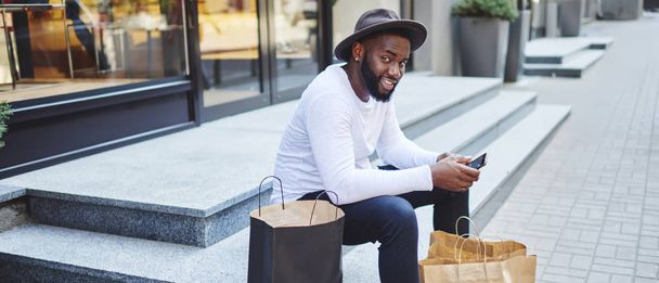 Retrato de los afroamericanos esperando los comentarios del servicio de tienda web sentado en las escaleras esperando la compra por teléfono inteligente, hombre oscuro positivo en el elegante sombrero descansado después del tiempo de compras. - Foto, imagen