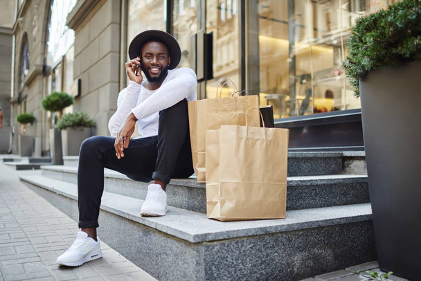 Happy afro american male buyer in trendy wear calling to friend for communicate about successful shopping, cheerful dark skinned guy having positive mobile conversation sitting on stairs with bags - Foto, immagini
