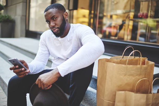 Portrait d'un afro-américain qui attend les commentaires d'un magasin en ligne alors qu'il est assis dans un escalier près d'un centre commercial, un homme à la peau foncée qui est un accro au téléphone intelligent et qui se repose après avoir fait ses courses - Photo, image