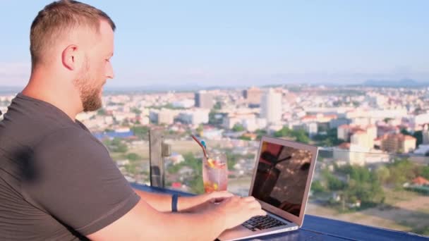 Mann arbeitet an einem Laptop in einem Café auf dem Dach mit schönem Panoramablick. Mann trinkt einen Cocktail und arbeitet am Computer. - Filmmaterial, Video