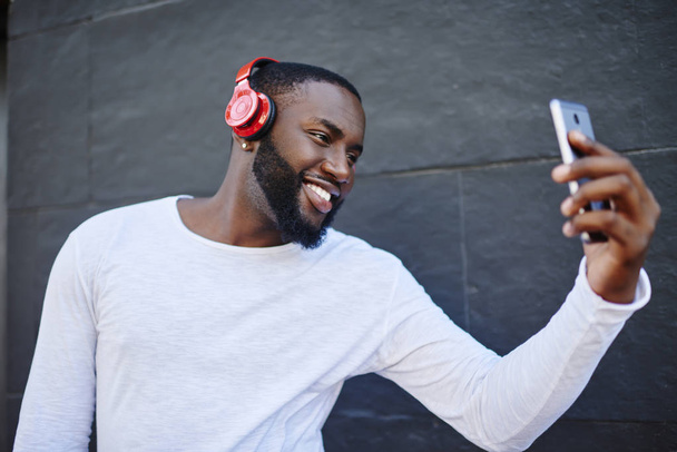 Cheerful man smiling at camera while making selfie on smartphone standing on urban setting and enjoying playlist via bluetooth headphones,positive blogger listening music in earphones and taking photo - Photo, Image