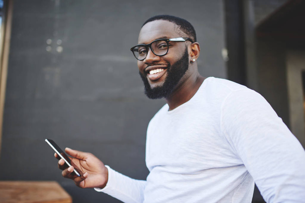 Happy smiling male blogger holding smartphone gadget in hand indoors and feeling good on leisure, portrait of cheerful hipster guy in eyewear with mobile phone spending time on publicity area - 写真・画像