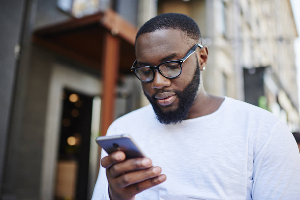 Millennial dark skinned man using smartphone app for booking online while spending time outdoors, african american hipster guy in optical spectacles read notification from chat on mobile phone - Фото, изображение