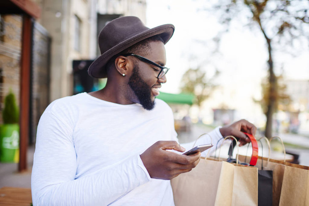 Smiling dark skinned hipster guy looking in shopping bags checking purchases satisfied with delivery booked via smartphone, cheerful african american male happy about online buying with discount - Foto, immagini