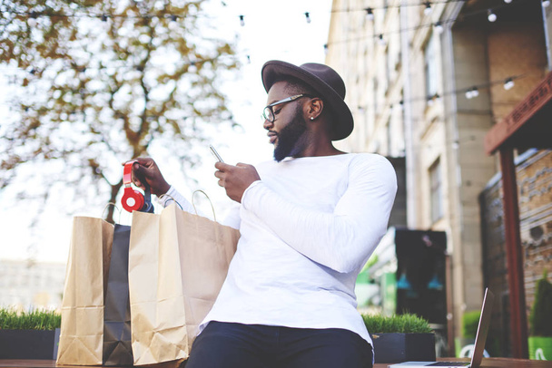 Dark skinned man checking purchases holding mobile phone and taking new bluetooth headphones for making picture and sending sms with photo to friend using public internet connection on urban setting - Foto, imagen