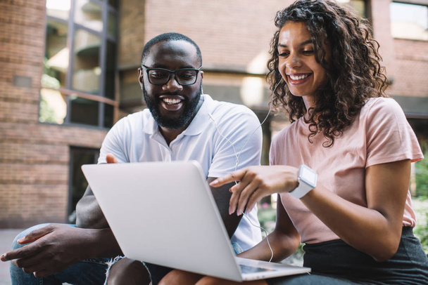 African American male and female bloggers taking rest togetherness laughing from funny content on website, positive woman with curly hair spending time with boyfriend pointing on laptop screen - Fotó, kép