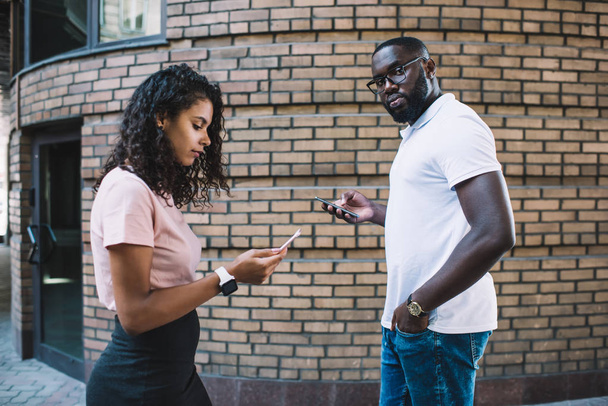 Male and female hipsters walking outdoors holding smartphones ignore real life, portrait of black man near concentrated girl on using mobile phone for chatting and networking addicted to gadget - Фото, изображение