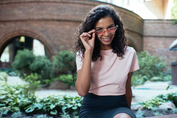 Half length portrait of cheerful female student dressed in stylish outfit resting outdoors during free leisure time, positive African American hipster girl in sunglasses - chanterelles posing - Photo, image