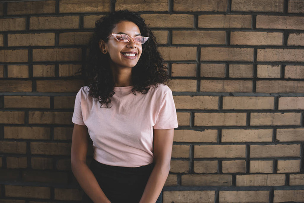 Excited African American female teenager in stylish pink sunglasses standing near promotional background, cheerful dark skinned hipster girl smiling near brick wall enjoying leisure and free day - 写真・画像