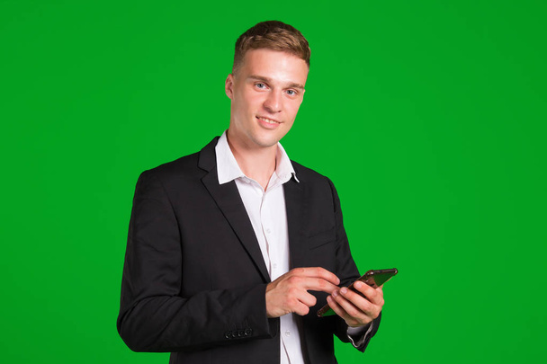Handsome man in a suit is using a smartphone and smiling, isolated on a green background. - Zdjęcie, obraz