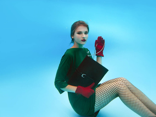 hermosa mujer joven bonita con estilo increíble en vestido verde, guantes rojos, bolso de embrague negro y medias en la red bajo el agua en la piscina. Concepto de moda
 - Foto, Imagen