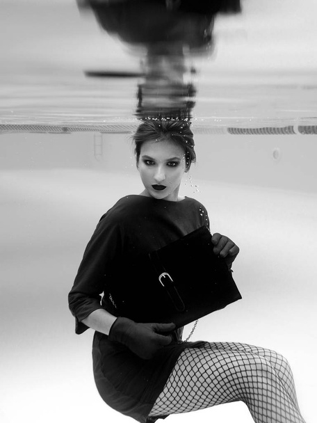 Monochrome beautiful amazing amazing pretty young woman in green dress, red gloves, black clutch bag and tights in the net underwater in the swimming pool. Концепция моды
 - Фото, изображение