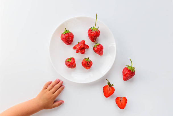 Strawberries in a plate on a white background and children's hands. Ugly food, top view, vertical orientation. - Photo, Image