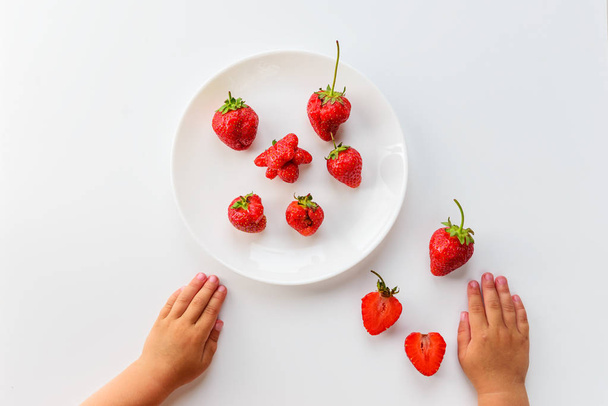 Fresas en un plato sobre un fondo blanco y las manos de los niños. Comida fea, vista superior, orientación vertical
. - Foto, Imagen