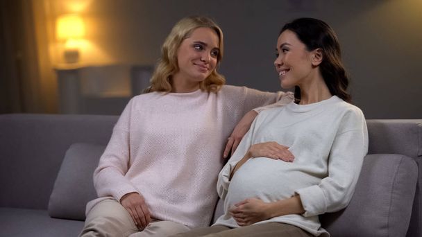 Young woman sitting on sofa with her pregnant friend, happy future mother - Fotoğraf, Görsel