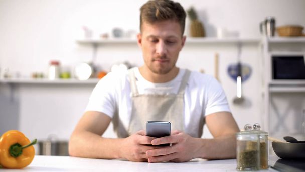 Jonge man in schort zoeken voedsel recept in mobiele telefoon, tips in culinaire blogs - Foto, afbeelding