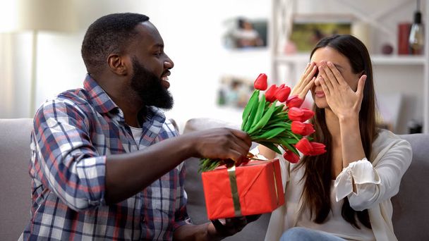 Black man presenting tulips and gift box to his girlfriend, holiday celebration - Φωτογραφία, εικόνα
