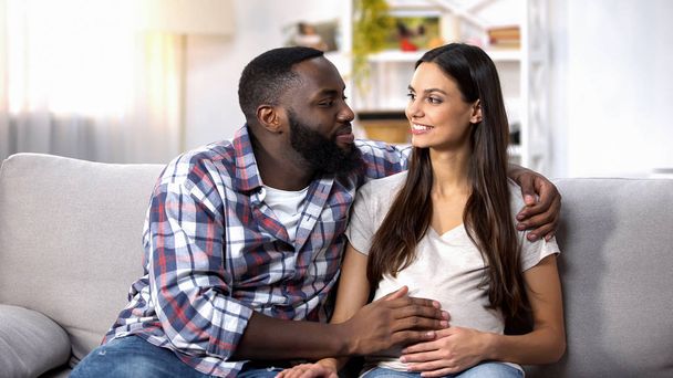 Cheerful Afro-American man stroking his pregnant wife belly, loving couple - Фото, изображение