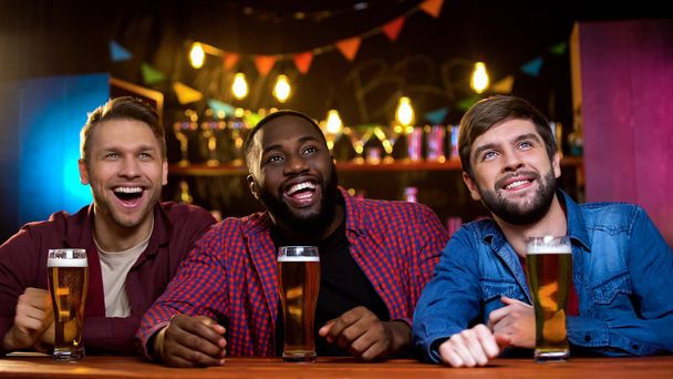 Multi-ethnic friends smiling watching comedy show on big screen in beer pub, fun - Foto, Imagen