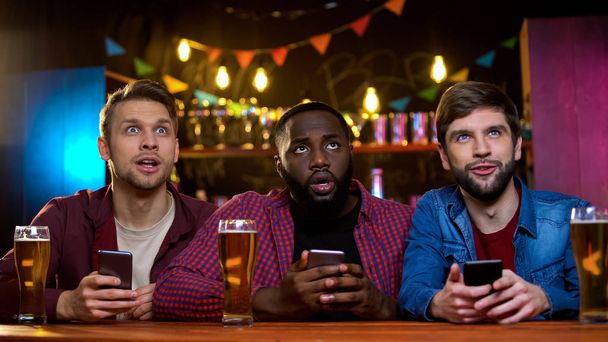 Mixed-race friends comparing lottery numbers on tv in bar with online tickets - 写真・画像