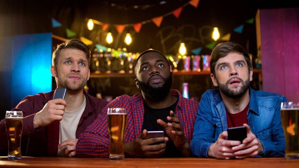 Multiethnic male friends watching football match in pub, upset with team losing - 写真・画像