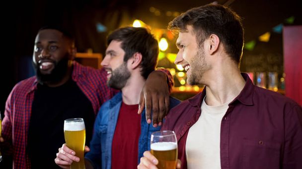 Caucasian friends looking at their african american friend, drinking beer in bar - Photo, Image