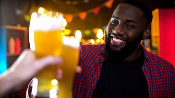 Cheerful african-american male clinking beer glass, enjoying results time in bar - Fotografie, Obrázek