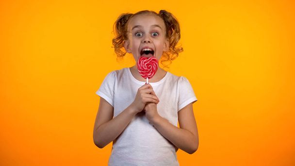 Pretty girl licking delicious heart-shaped lollipop, childhood, confectionery - Foto, Bild