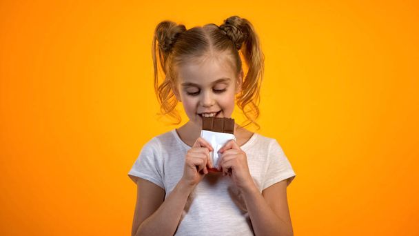 Rozkošná holčička, která kouše lahodnou čokoládu požívající sladkou chuť šťastné dítě - Fotografie, Obrázek
