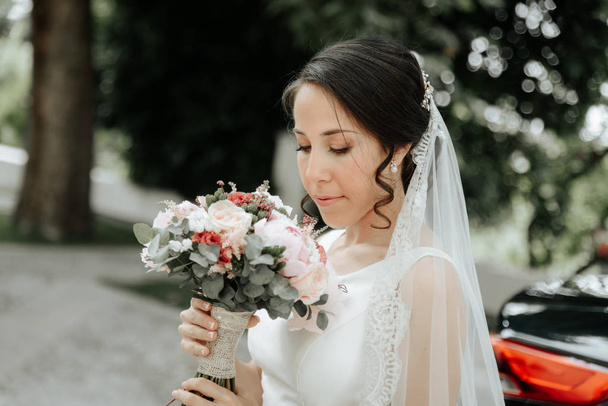 Novia sonriente en un vestido de novia y velo, mira el ramo de bodas
. - Foto, imagen