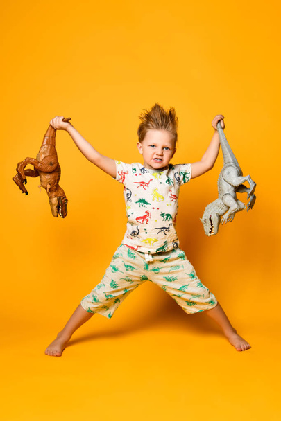 piccolo ragazzo allegro tiene un dinosauro di plastica in ogni mano. Il ragazzo è vestito con un vestito con immagini di dinosauri. - Foto, immagini