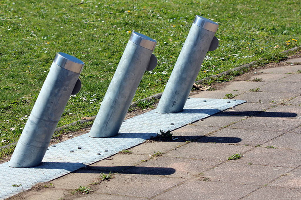 Sıcak güneşli bahar gününde taş karolar ve kesilmemiş çimlerle çevrili metal standa monte edilmiş kiralık elektrikli bisikletlerin park edilmesi ve şarj edilmesi için kullanılan eğimli metal direkler - Fotoğraf, Görsel