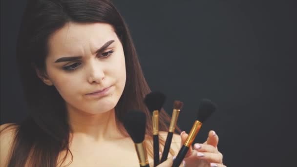 Young pretty cute girl standing on a black background. At the same time it holds in the hand brushes for make-up choosing the one that is needed. Has a pretty clean skin. - Кадры, видео