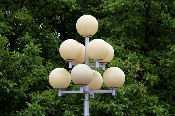 Vintage retro multi level pyramid style street lights with multiple ball shaped lights mounted on top of strong metal pole with dense trees in background on warm sunny spring day - Photo, Image