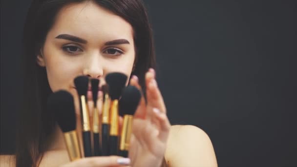 Young pretty young girl posing with makeup on black background. Holds a brush in your hands and chooses the right brush. - Кадры, видео